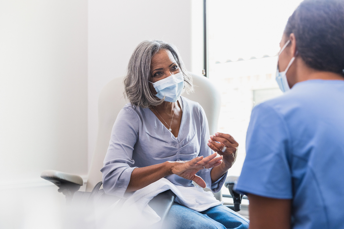 A senior woman, wearing a protective face mask, talks with a female nurse during a medical appointment.