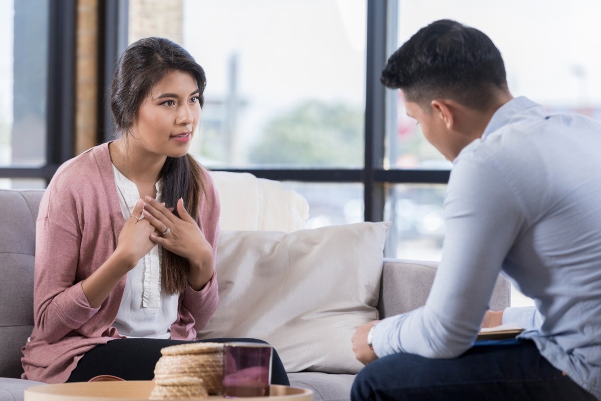 serious young woman sits on a couch across from an unrecognizable counselor and gestures as she speaks to him.