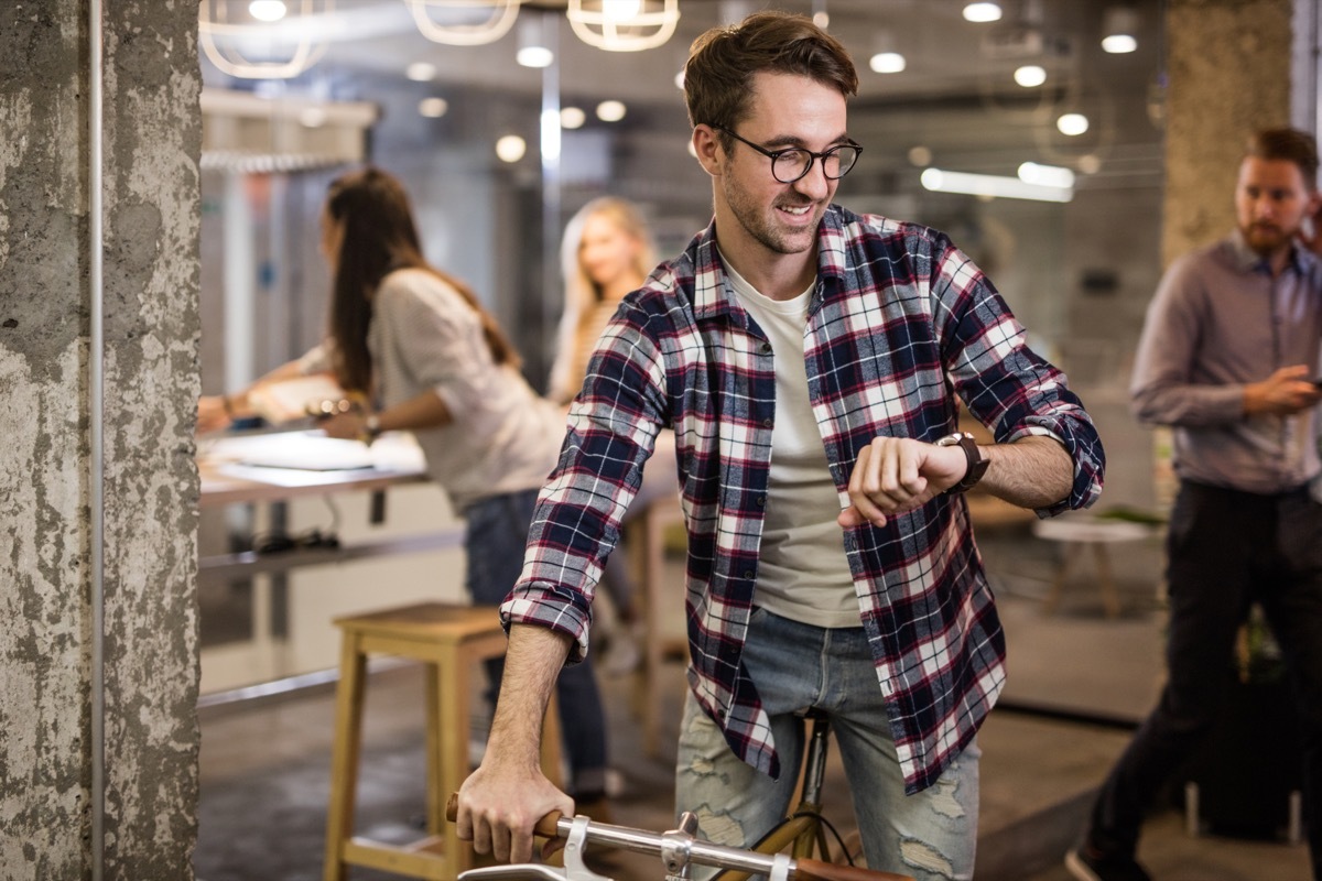 Happy creative man checking the time while being on a bike at casual office. There are people in the background.