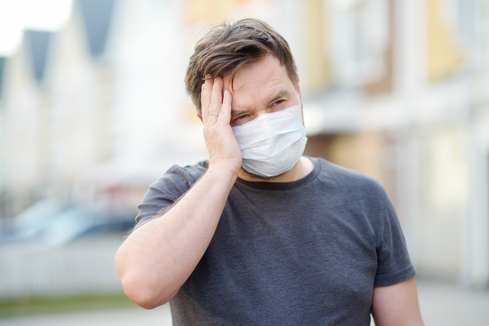 A middle-aged man wearing a face mask holds his head in pain, perhaps with symptoms of COVID-19.