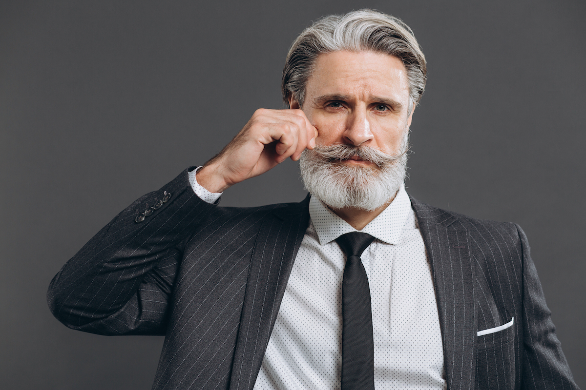 Man wearing a dark gray suit with gray hair, beard, and curly mustache