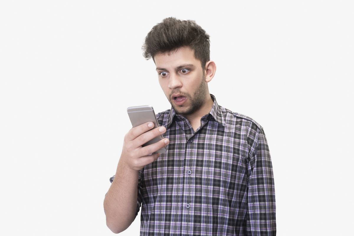 Close-up portrait of shocked young man looking at smart phone's screen receiving bad news on a white background