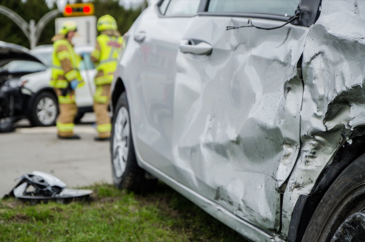 Close up on a car crashed during an accident with two firemen in background with firetruck during a day of summer