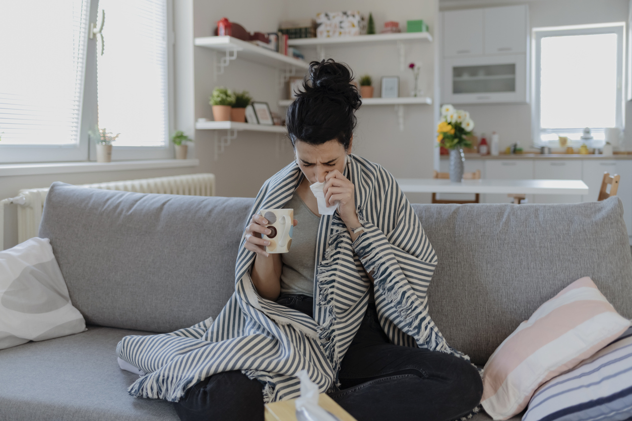 A young woman sits on a couch wrapped in a blanket with COVID symptoms