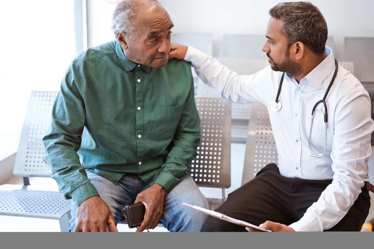 Healthcare worker giving bad news with hand on shoulder of male patient. Doctor consoling sad senior man in waiting room. They are sitting at hospital.