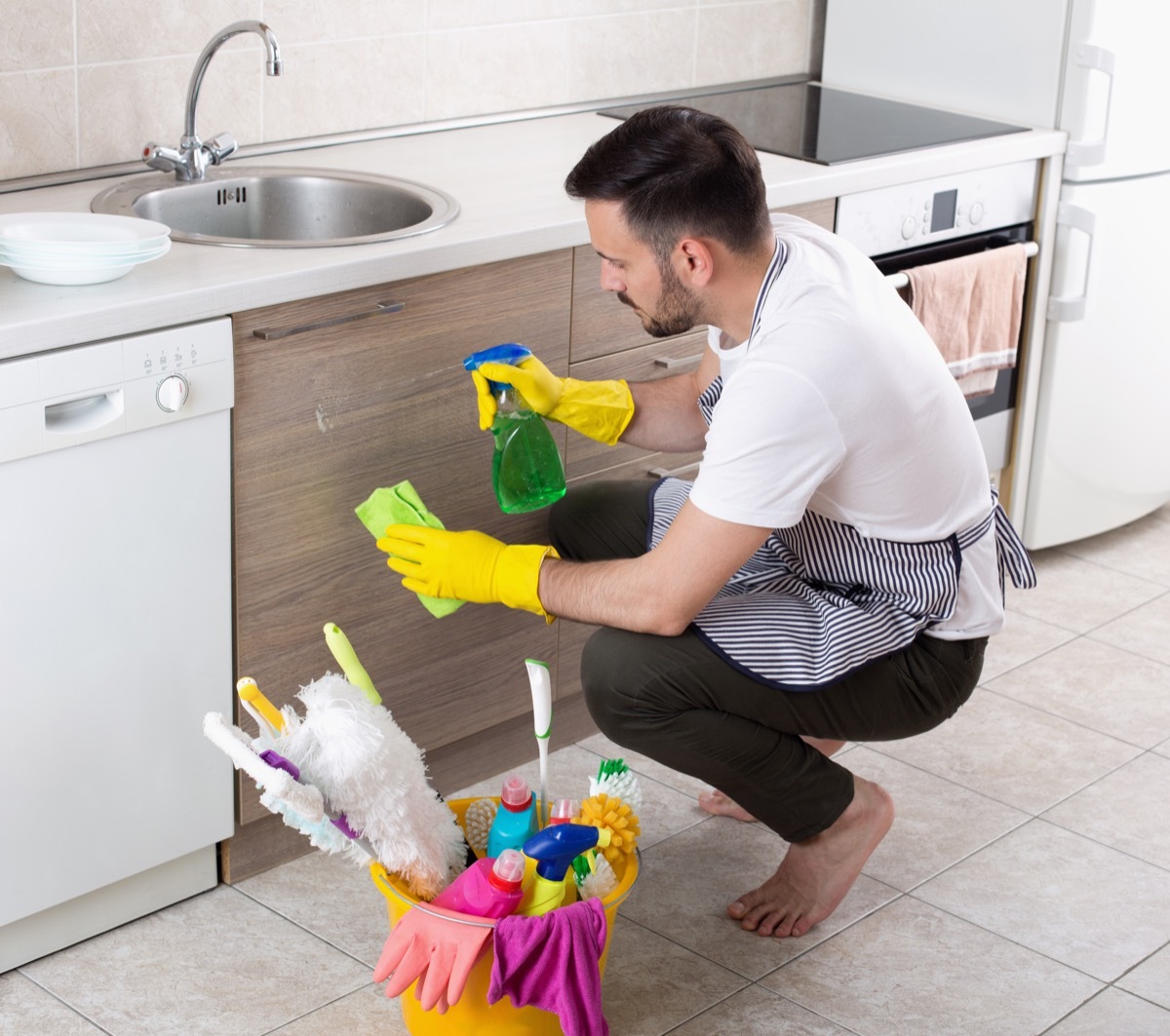 Handsome young man with apron and protective gloves wiping kitchen cabinets. House cleaning service