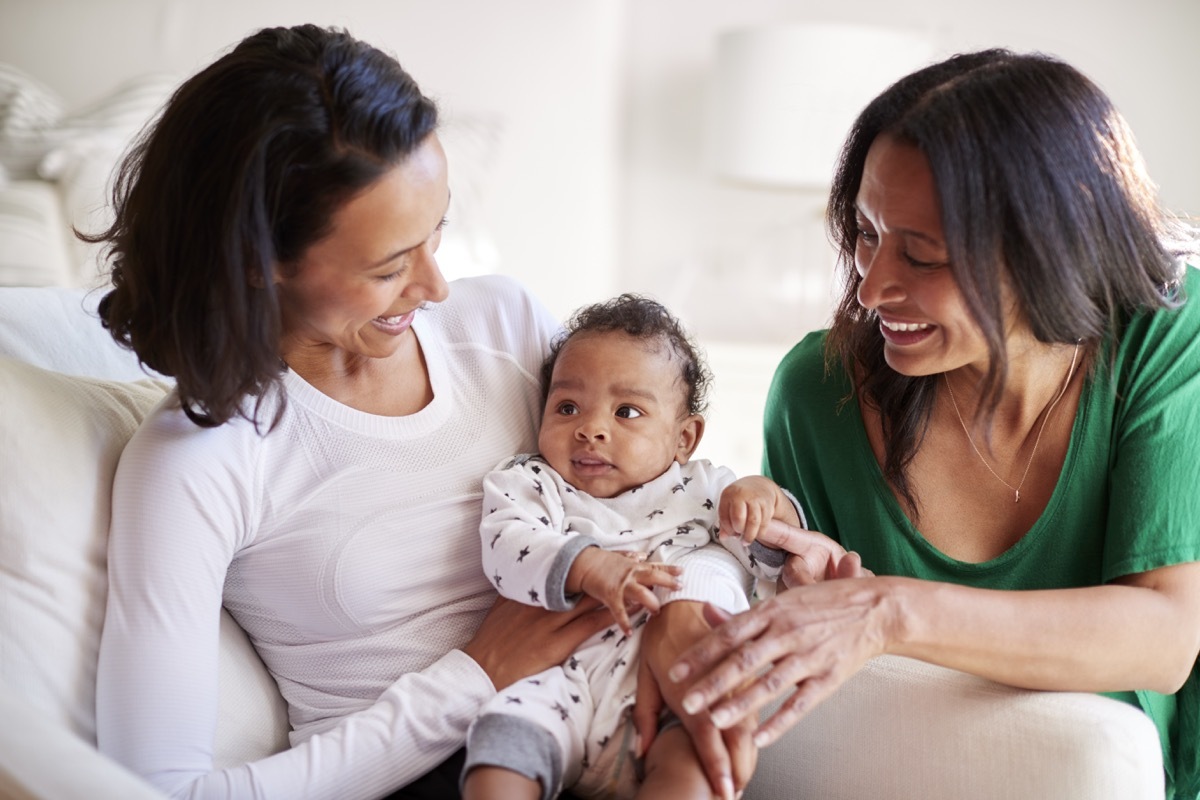 Millennial mother sitting in an armchair holding her three month old baby son, her mother kneeling beside them