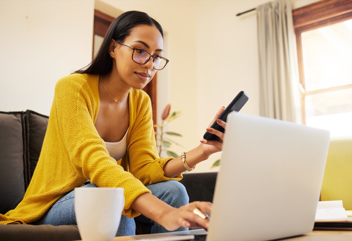 Woman working remote while typing on her laptop and holding her smartphone sitting on a sofa in a bright living room