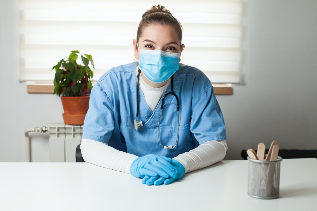 General Practitioner sitting by her desk in office,wearing blue uniform,protective gloves & face mask,virtual tele visit via video call