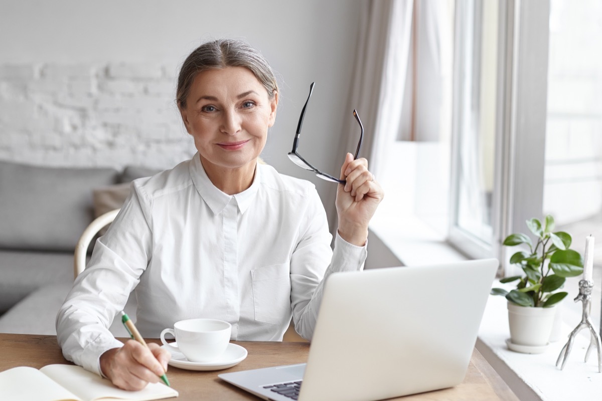 mature female working in modern office, holding eyeglasses and writing down in a notebook with her laptop open