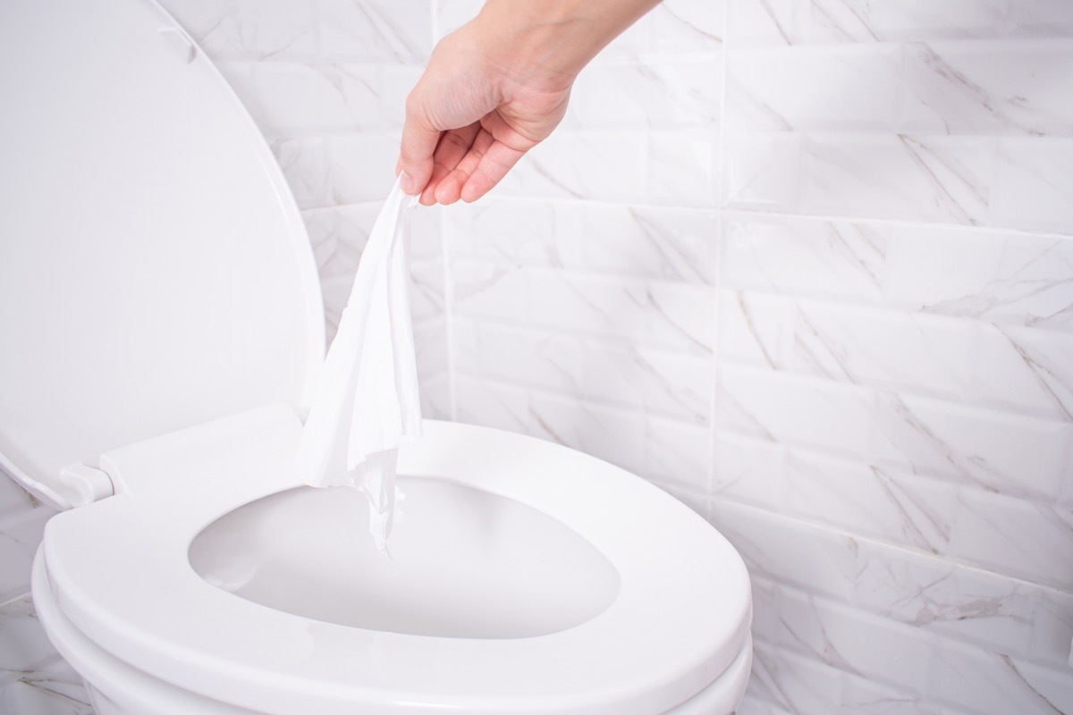 Close up hand throwing toilet paper to the toilet in a white tile bathroom.