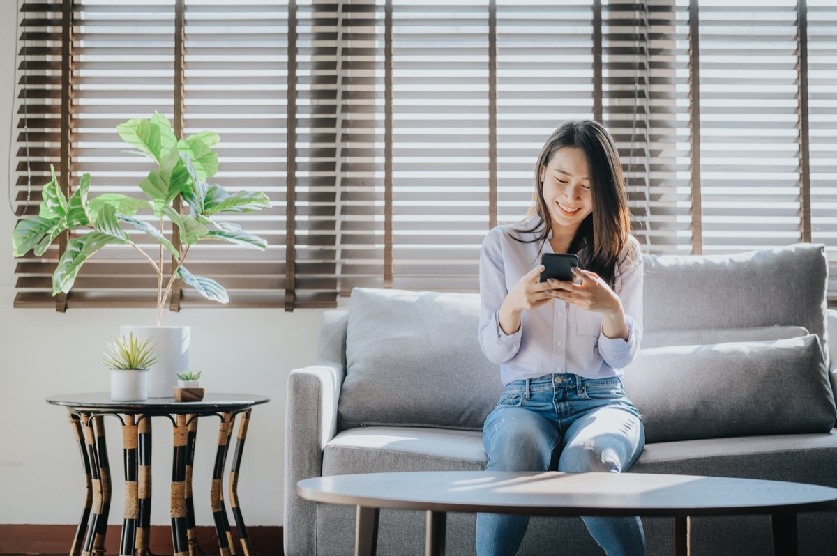 Happy woman smiling and texting on the smartphone sitting on sofa i lving room at her home