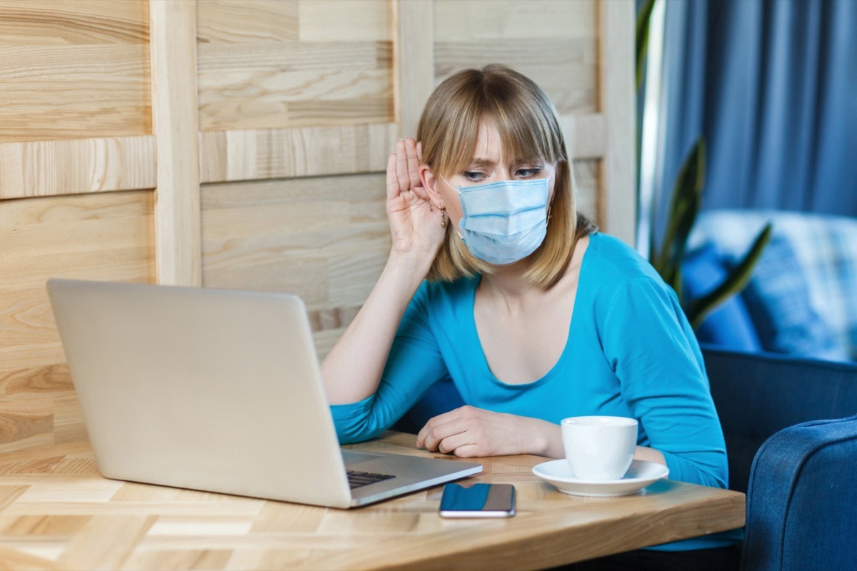 woman with surgical medical mask is sitting and working on laptop and trying to hear with attention to laptop