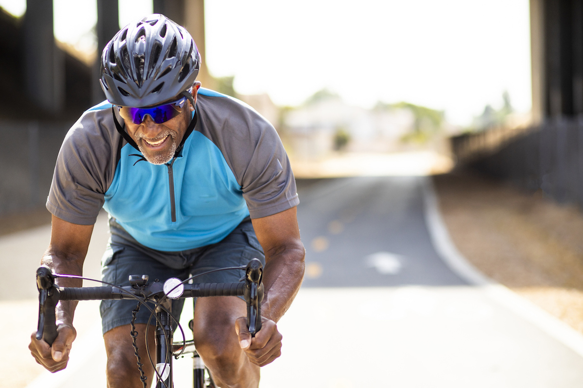 A senior black man sprints on his road bike training for a race. He's smiling and wearing a helmet.