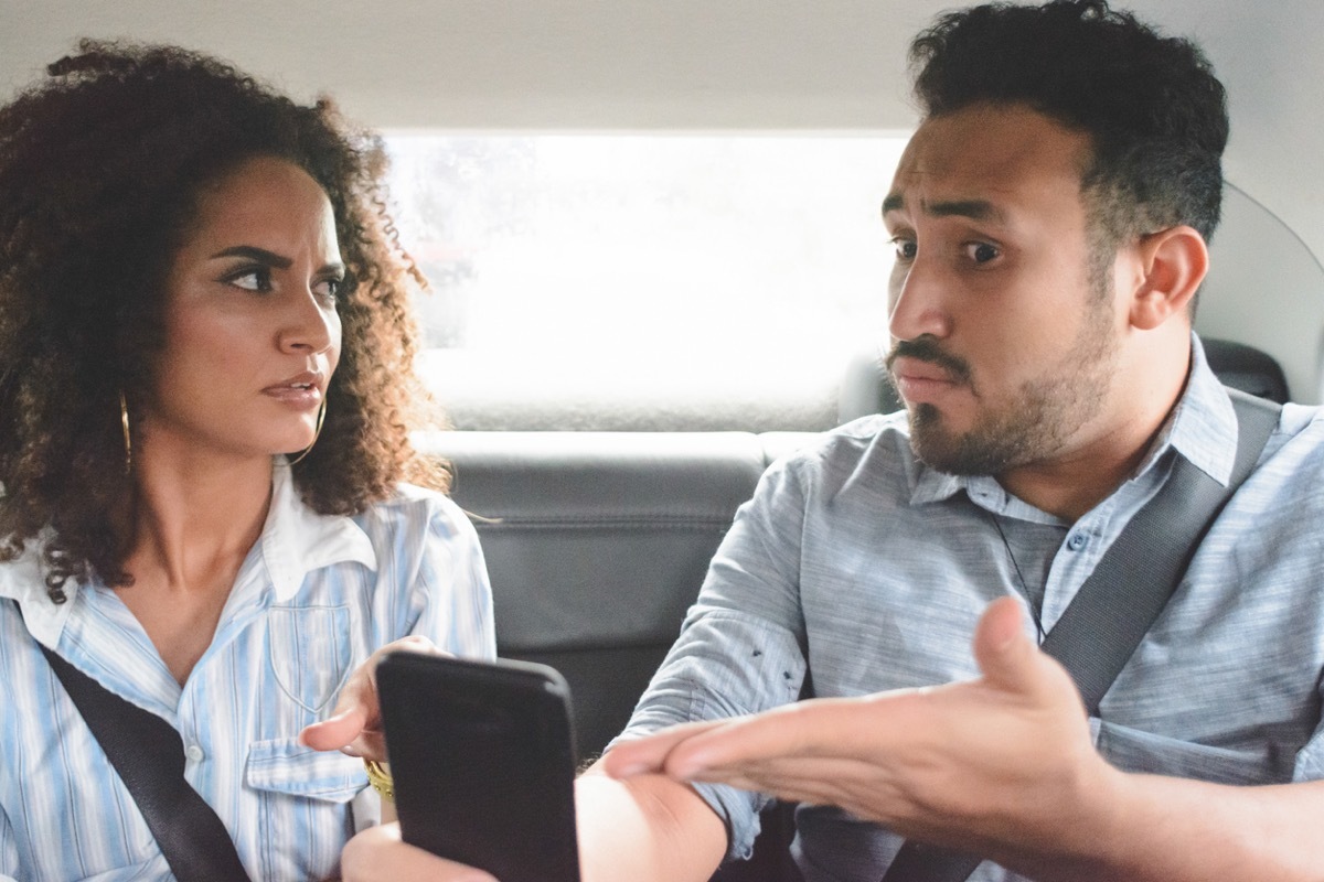 young couple arguing in the backseat of a car while the man points to his phone defensively