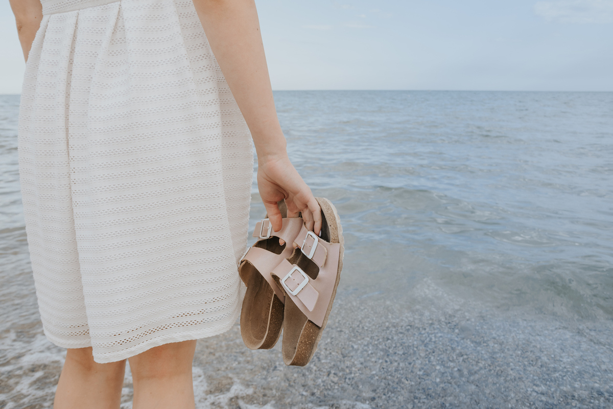 Woman in an off-white dress stands near the ocean holding beige Birkenstock sandals