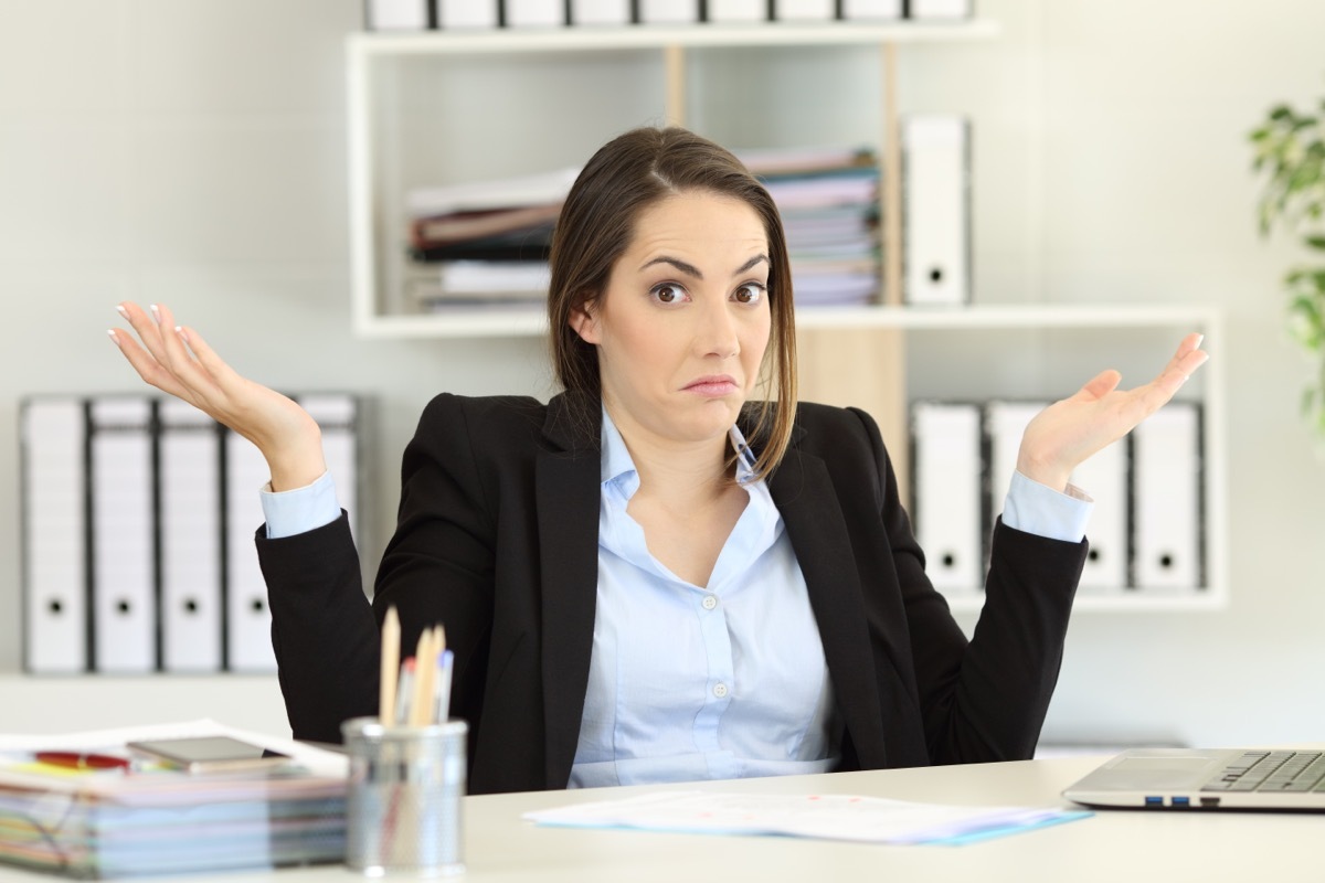 A young woman wearing a shirt and blazer shrugs while sitting at her office desk