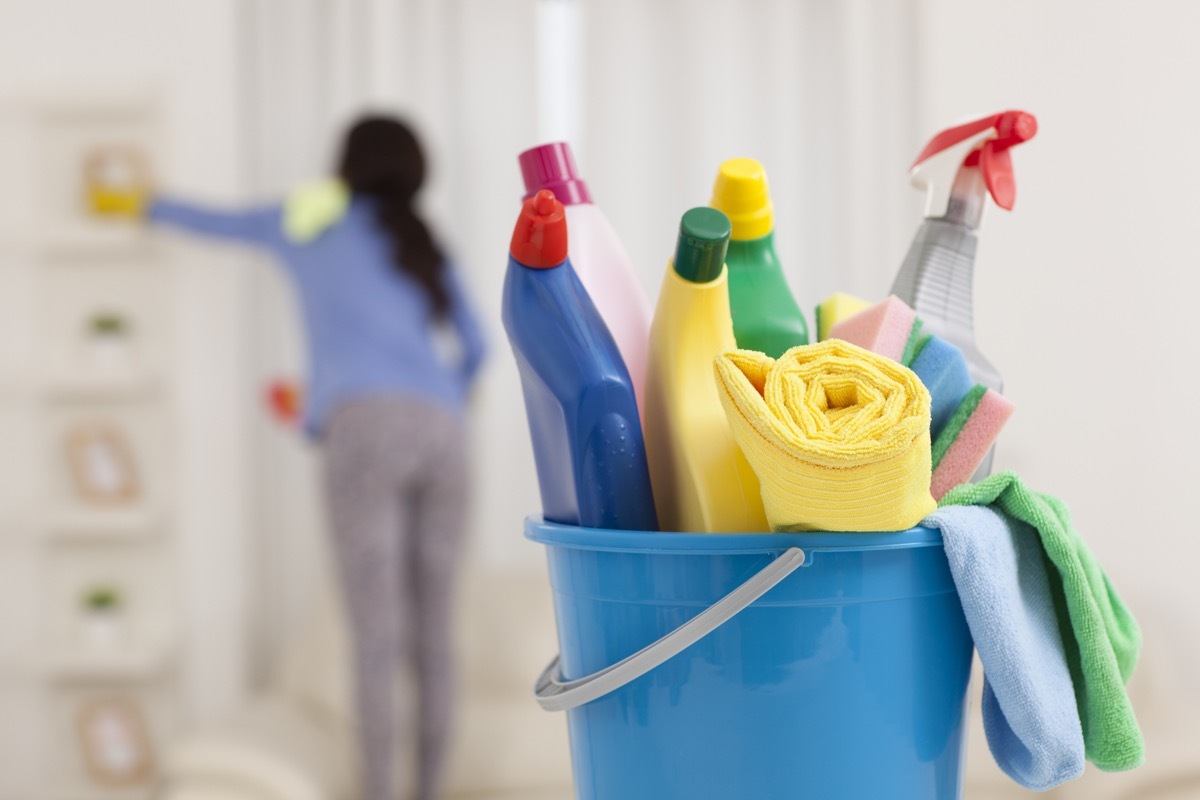 Group of home cleaning products on foreground and rear view of unrecognised woman cleaning the shelves in the living room.