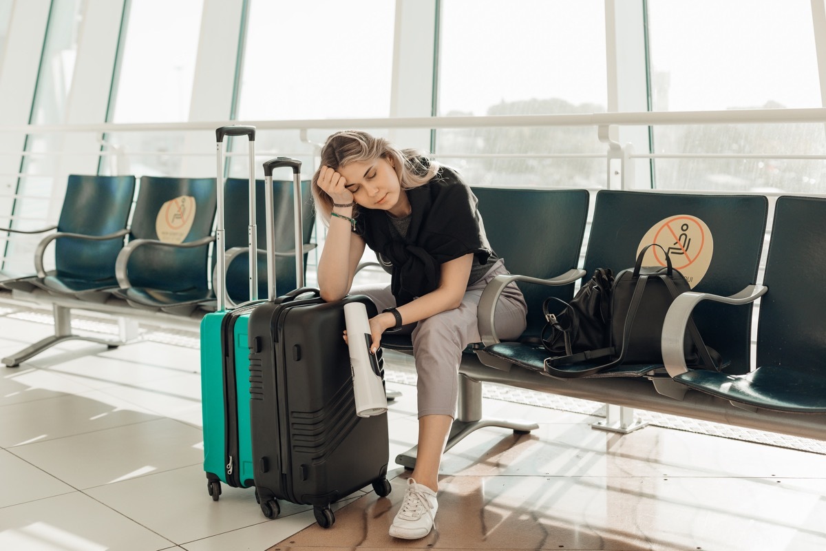 Bored blond woman with luggage, leaning elbow on bags, sitting in waiting room at airport due to coronavirus pandemic Covid-19 outbreak travel restrictions. Flight cancellation. Too late for voyage