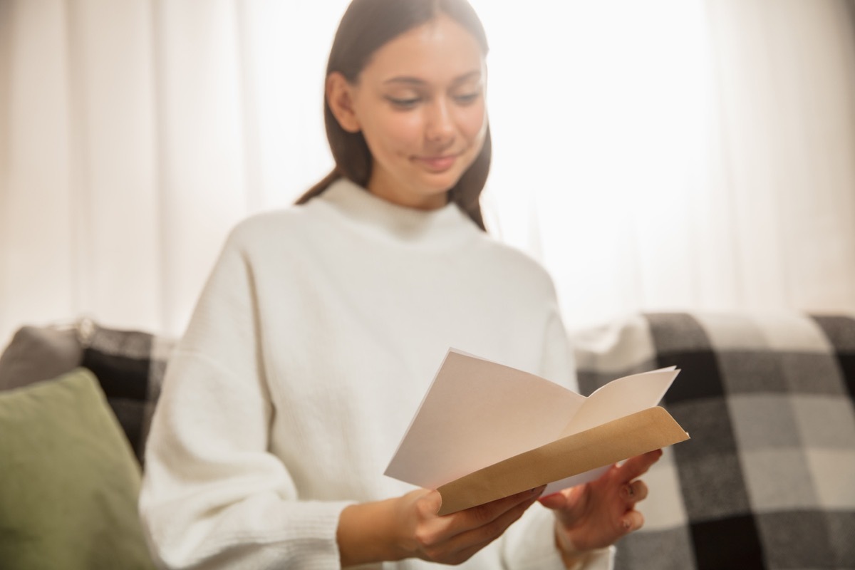 Woman opening, recieving greeting card for New Year and Christmas 2021 from friends or family. Reading a letter with best wishes, looks happy, cheerful, smiling, opening envelope. Holidays, celebration.