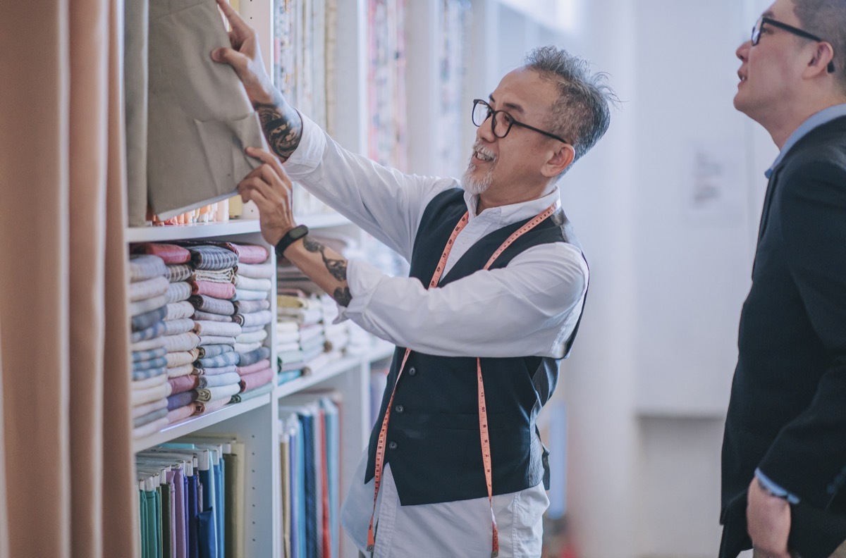 enior man tailor picking and showing his customer on fabric garment choice on new tailored clothing suggestion at his store shelf display