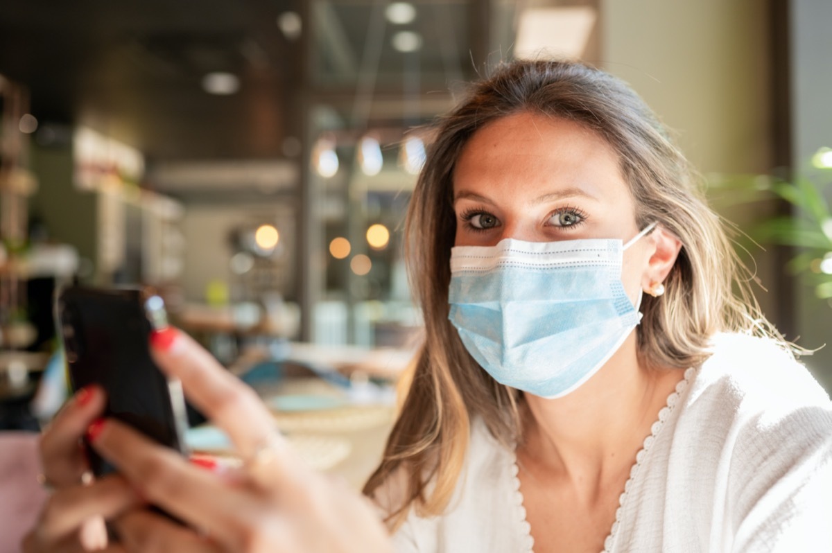 Woman in a face mask sitting at a bar, looking at the camera while holding her smart phone in her hands