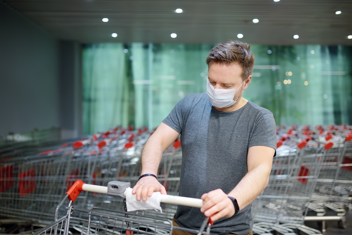 Man wearing disposable medical face mask wipes the shopping cart handle with a disinfecting cloth in grocery store