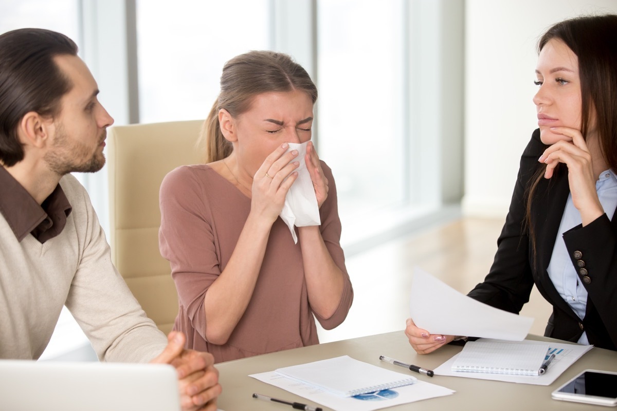 woman with handkerchief has sneezing attack, blowing nose while working with colleagues on meeting, caught cold, flu symptom