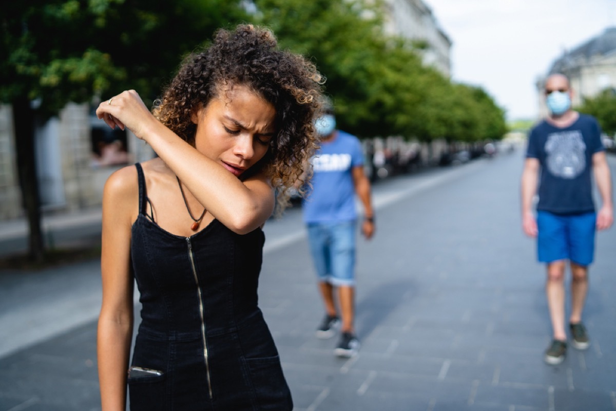 young black woman coughing into her arm outside with people in masks behind her