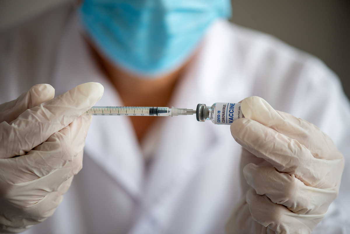 Hand with white surgical gloves taking coronavirus vaccine dose from vial with syringe