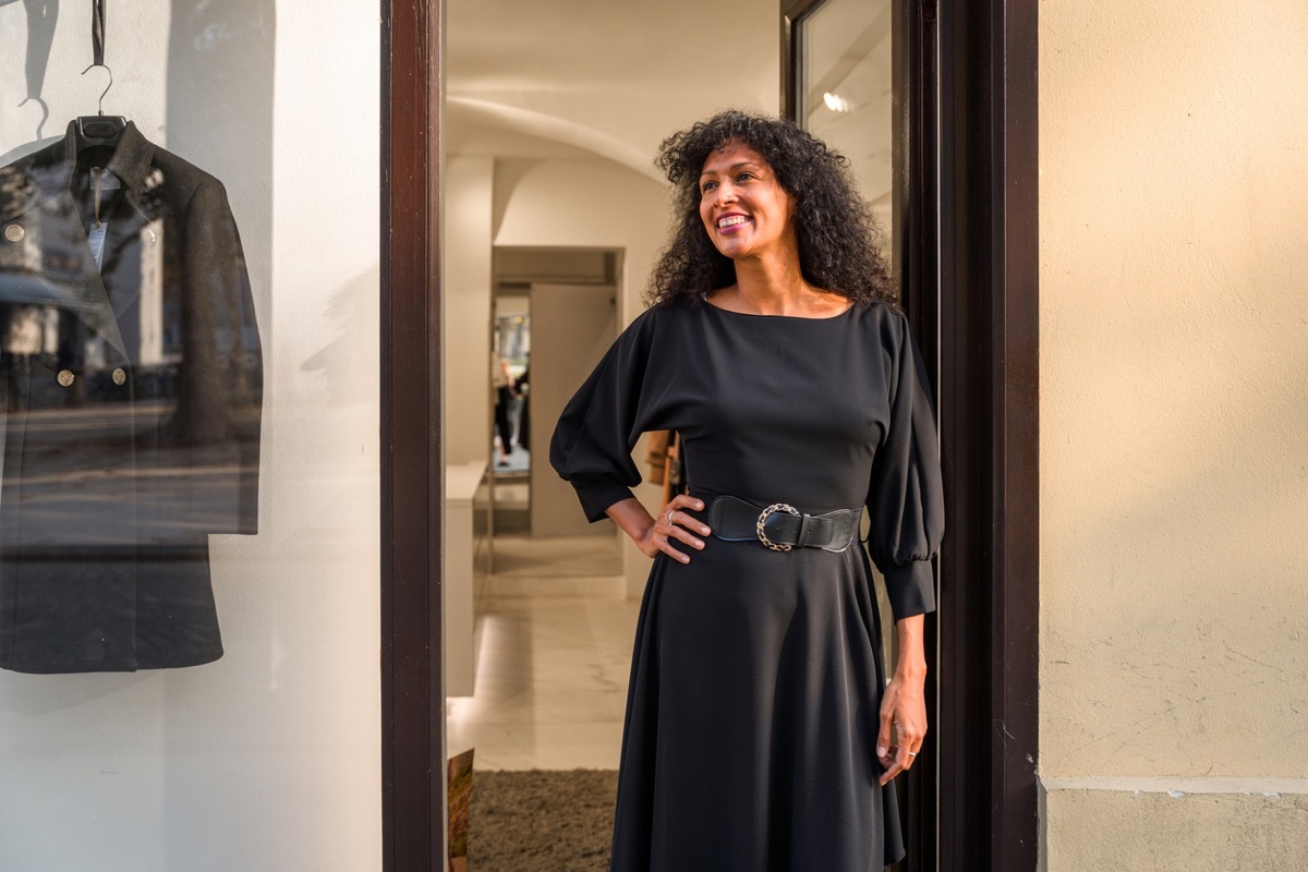 A beautiful and elegant female model posing in front of a classy fashion shop. Wearing a light black dress with a belt, her black hair down. Looking away, 3/4 length image.