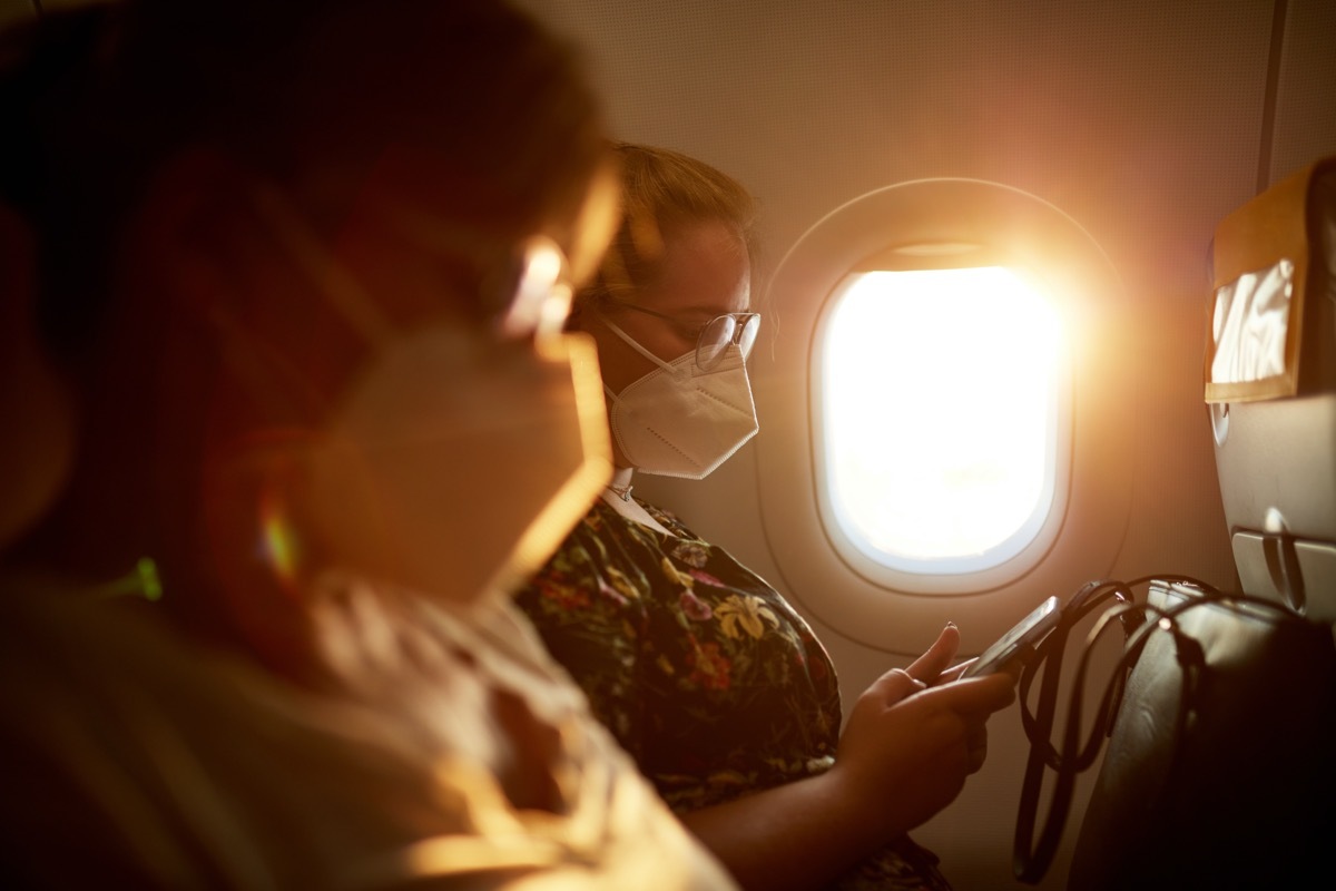 Side view with focus on background of mature Caucasian mother and teenage daughter wearing protective masks as they travel by airplane in time of COVID-19.