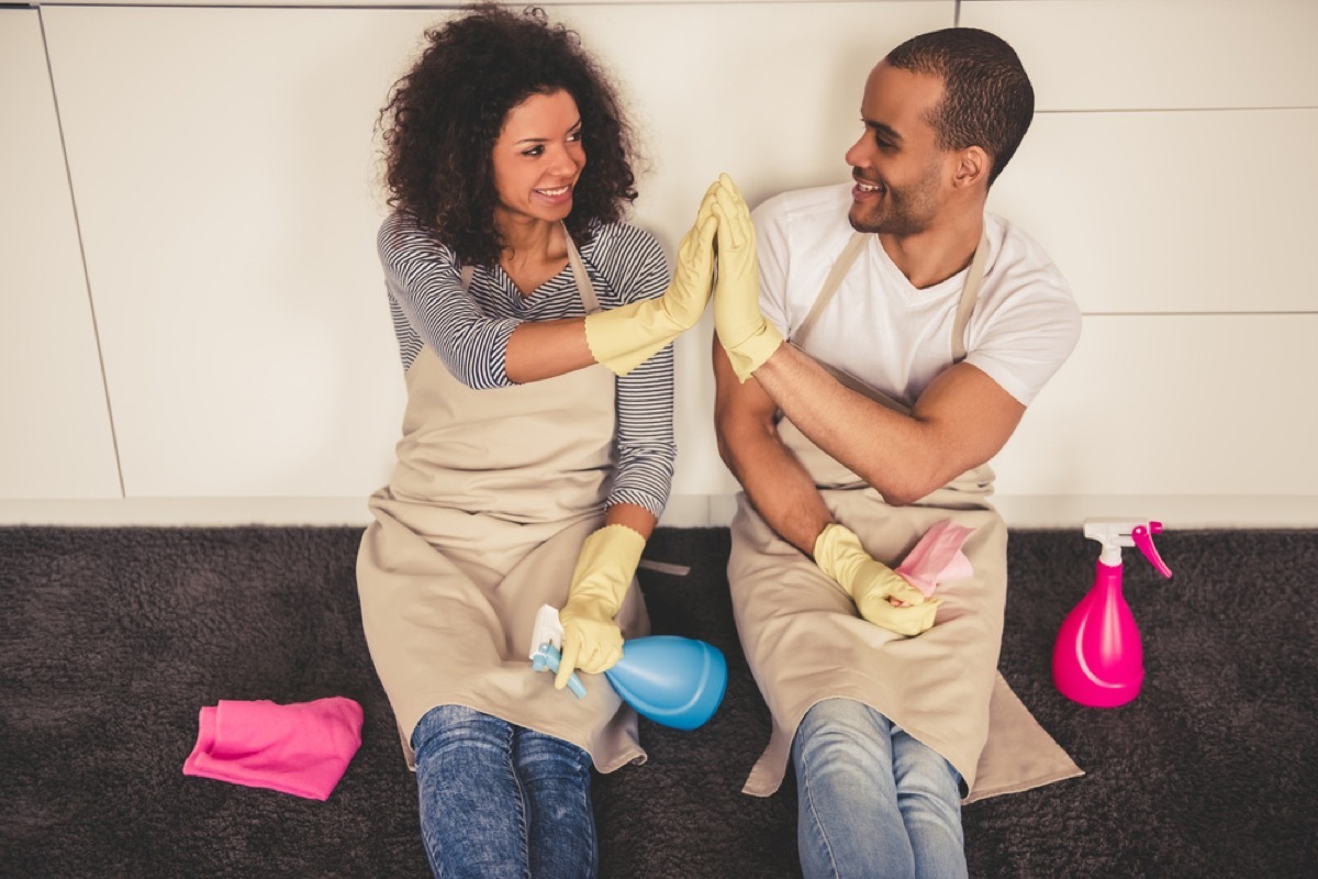 young black couple wearing cleaning gloves high fiving while sitting on kitchen floor with cleaning products