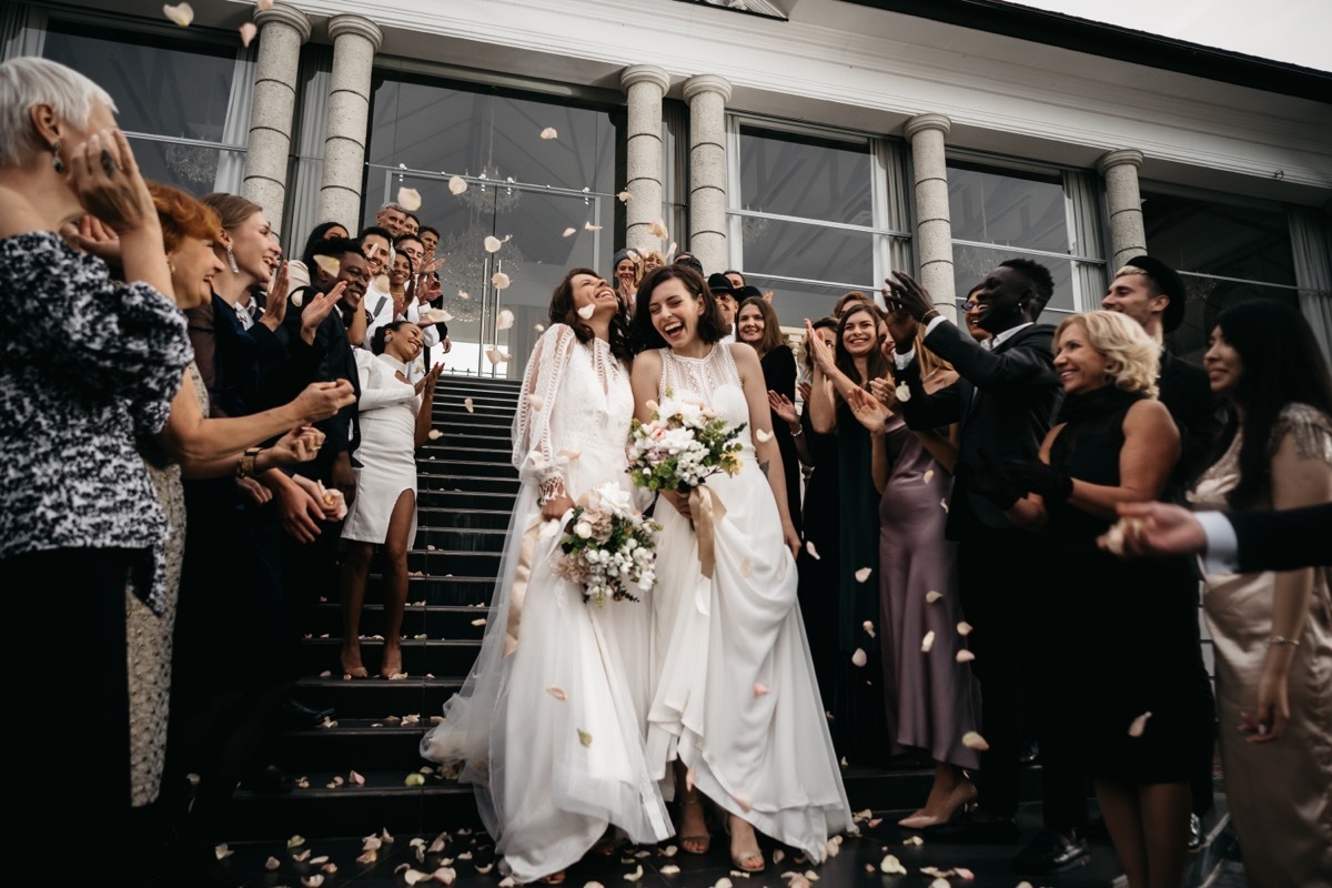 Two brides walking down steps, being showered with rose petals by wedding guests