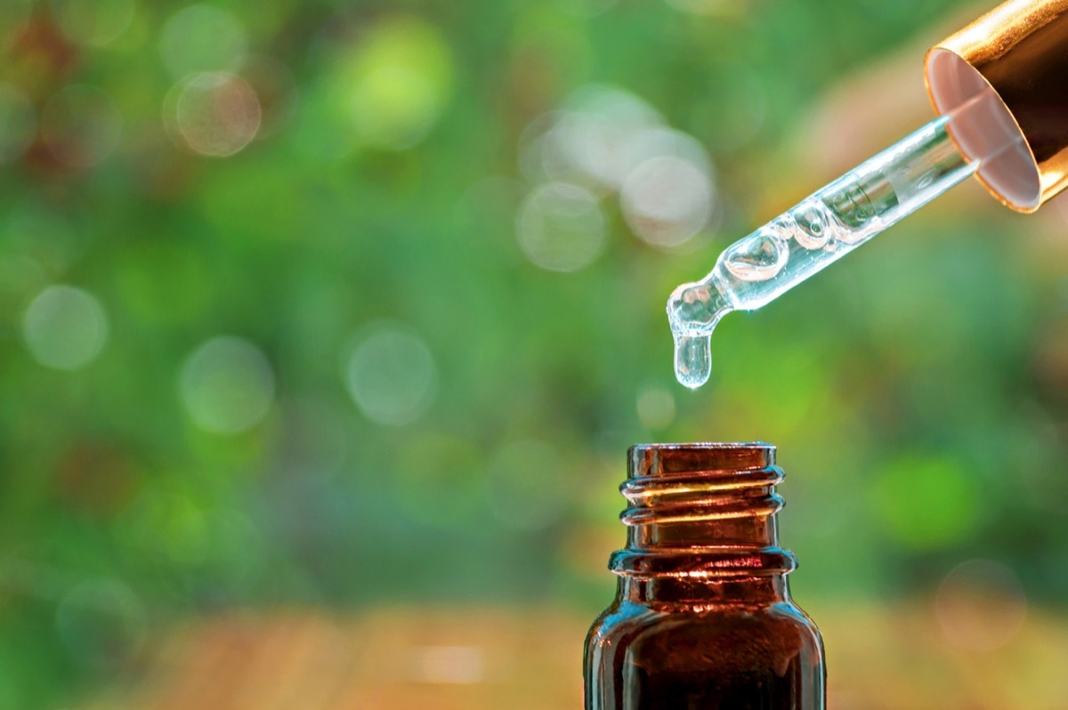 Pipette with essence drop and bottle, closeup on blurred nature background. Herbal essential massage oil dripping into bottle. Spa beauty concept. Selective focus.