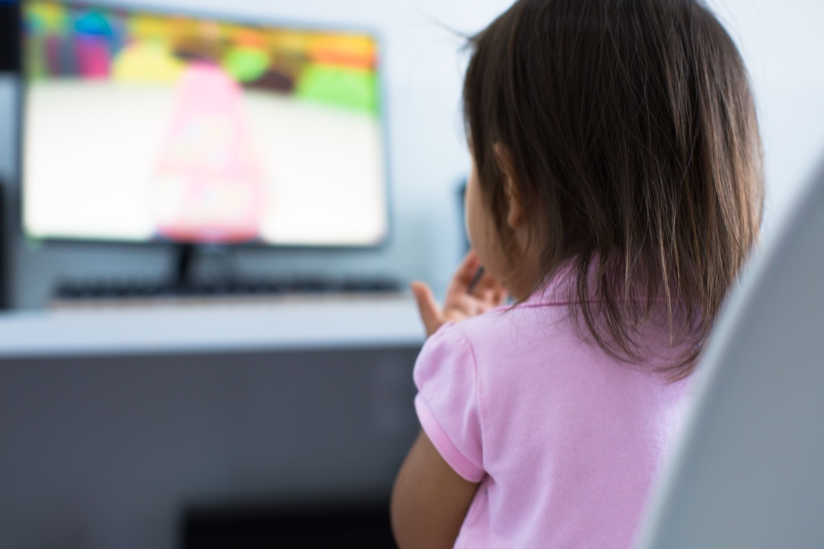 A 3-5 years old girl toddler sitting at the computer desk watching cartoon shows on the monitor at home. Children educational and development concept.