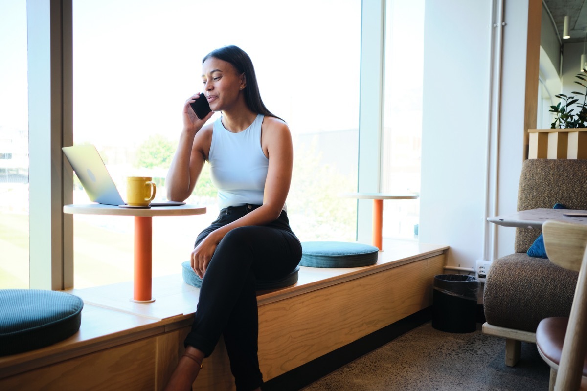 Smiling young businesswoman sitting in a table in an office lounge and talking on her phone while working on a laptop