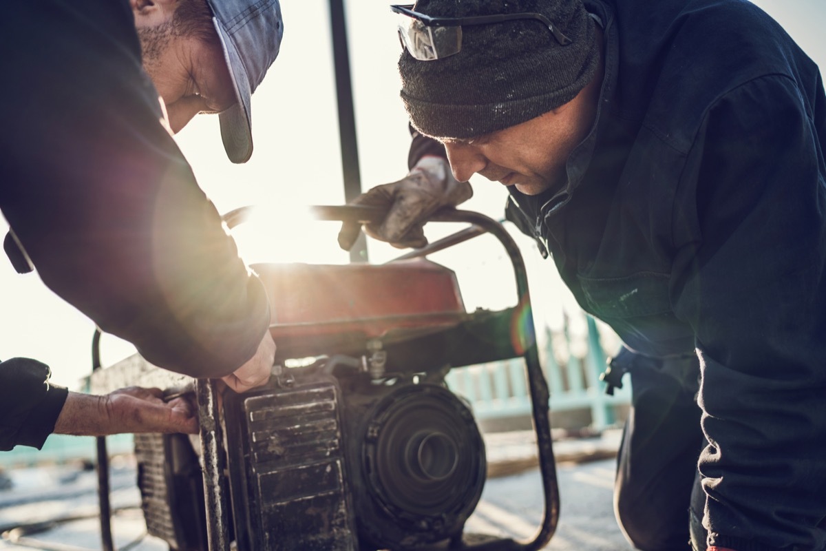 Two construction workers repairing power generator outdoors.