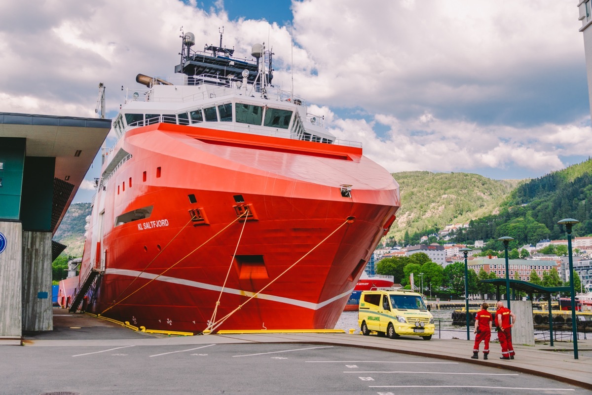 Two Norwegian paramedics in red uniforms are resting near an ambulance parked in a port near a large ship. Theme healthcare and medicine in Norway, Bergen July 28, 2019.