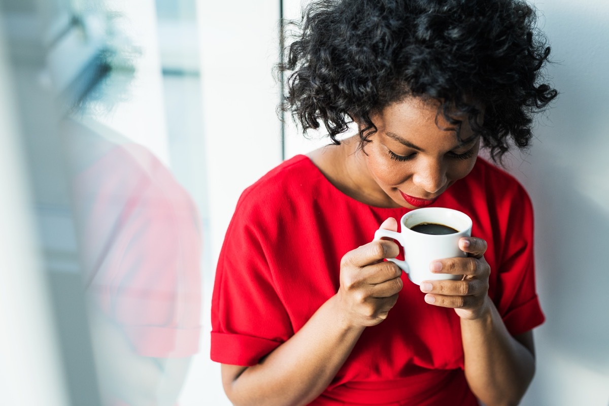 A close-up of a woman standing by the window holding a cup of coffee, smelling it. Copy space.