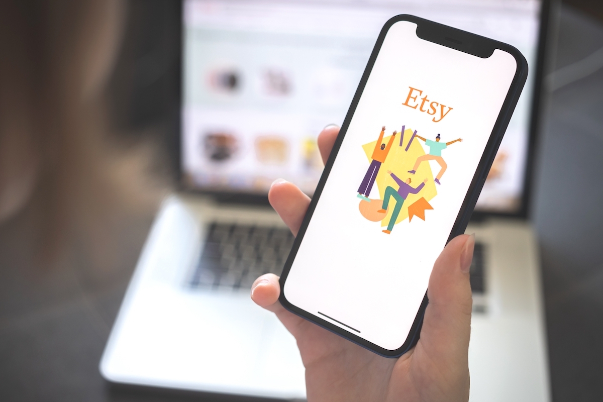 Hand holding a phone with the Etsy app, with a blurred laptop in the background