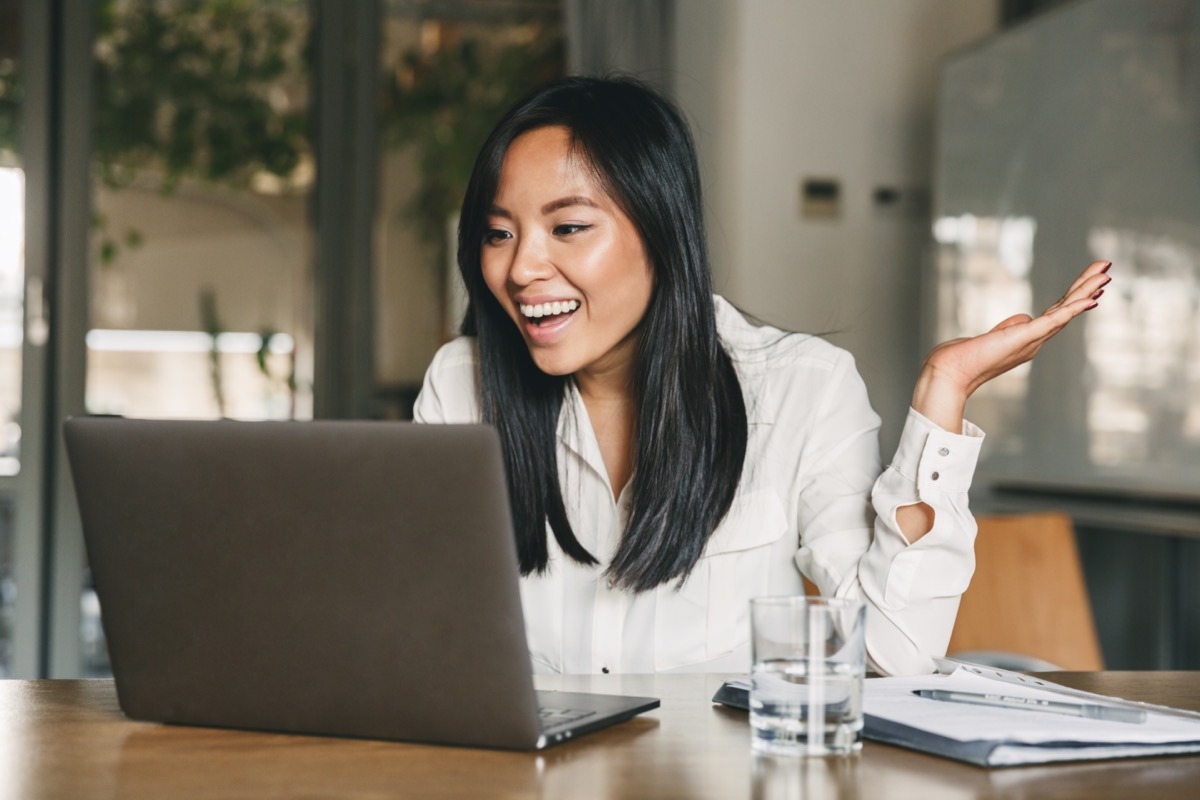 woman 20s wearing white shirt smiling and gesturing hand aside while speaking or chatting on video call at laptop in office