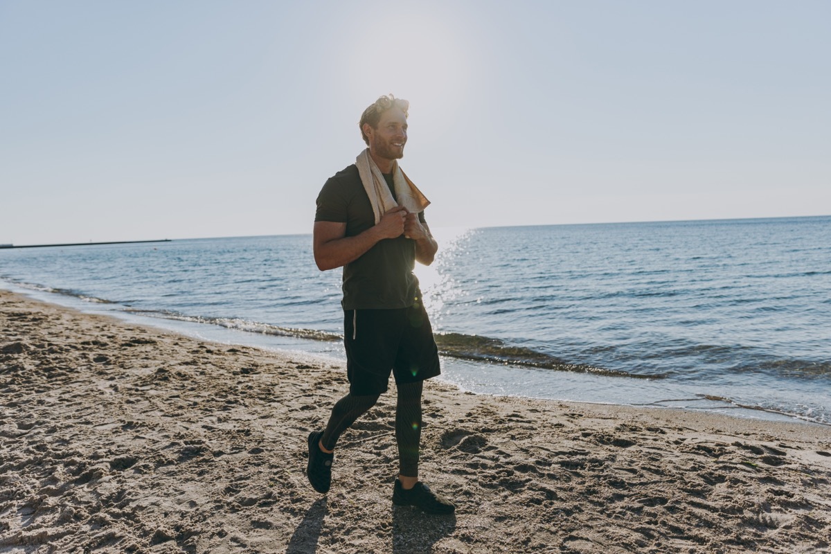 Full size happy young strong athletic toned fit sportsman man in sports clothes towel warm up training walking look aside at sunrise sun dawn over sea sand beach outdoor seaside in summer day morning.