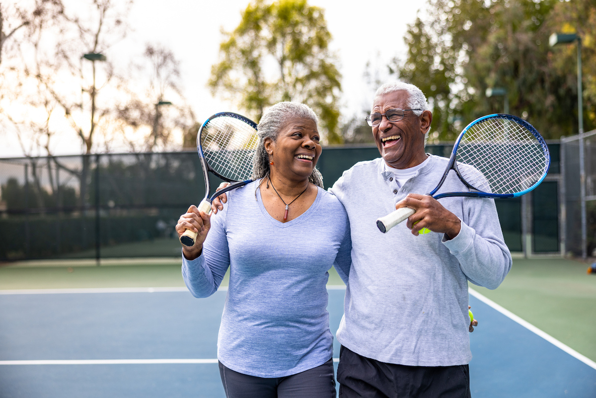 A senior black couple laughing and smiling, leaving the tennis court after their workout.