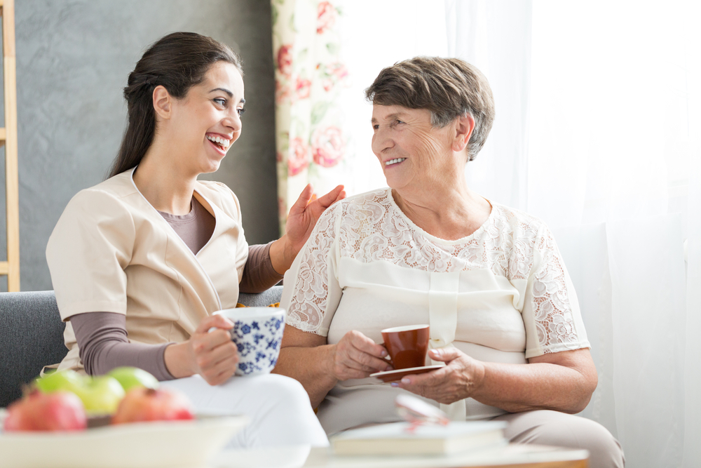 A young nurse sits next to a senior woman on the couch while drinking out of mugs and smiling