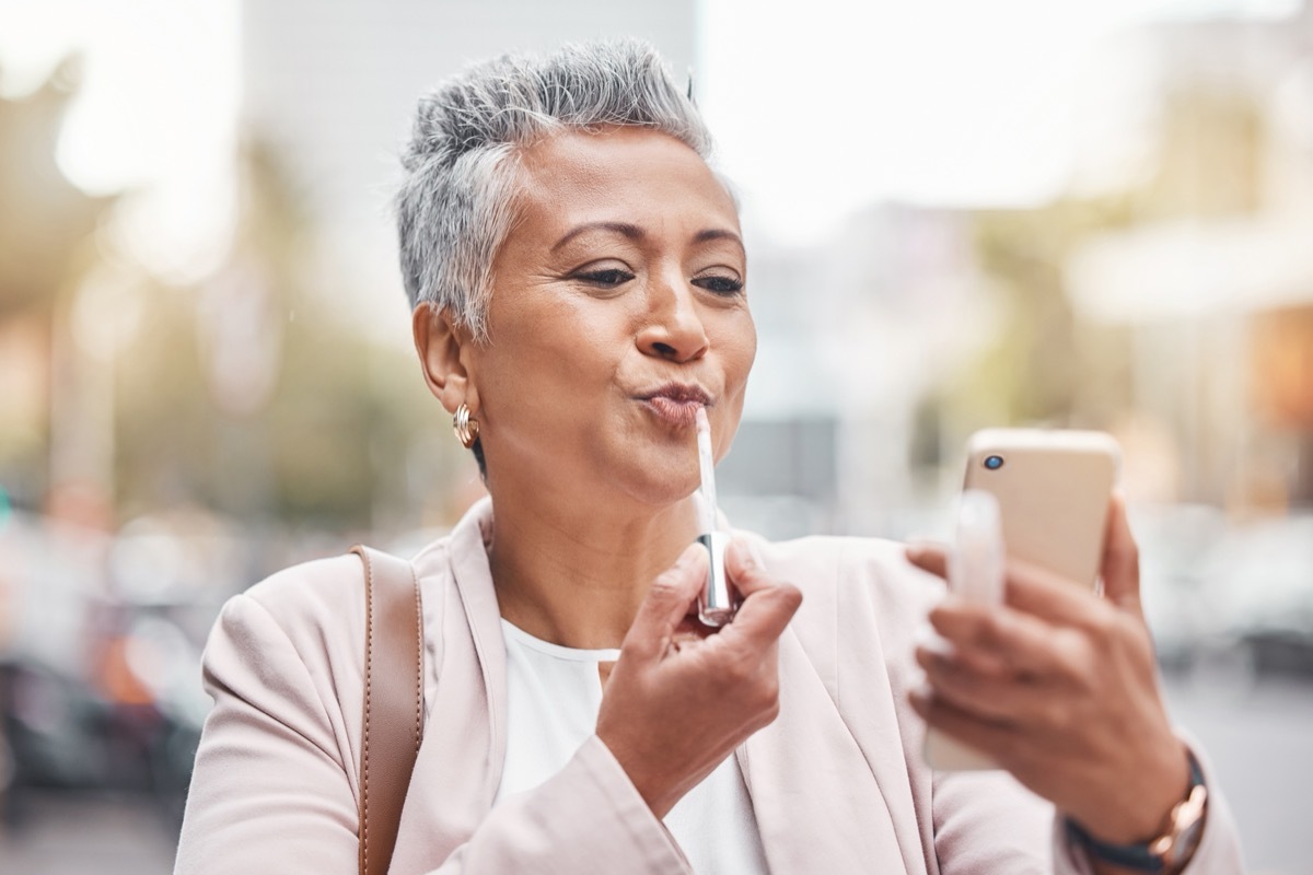 Woman, phone and lipstick in city for makeup, happy or cosmetics for financial service career. Accountant, cosmetic and face beauty by blurred background in metro, summer and smartphone for aesthetic