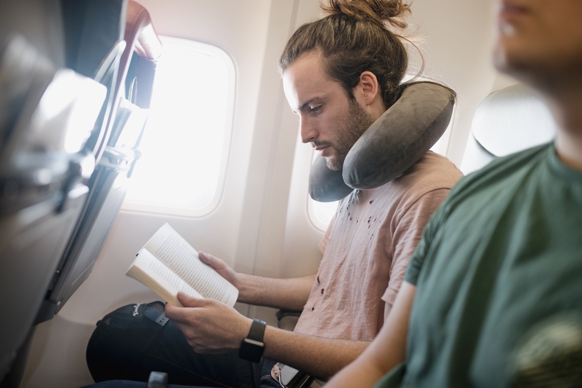 One young man is reading a book while travelling on an airplane. He is using a neck pillow.