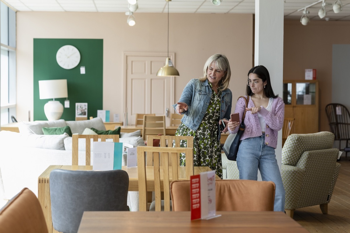 A mature woman and her daughter looking around a furniture store together for new furniture for home. They are looking for an item that the daughter has up on her phone, that she has seen online.