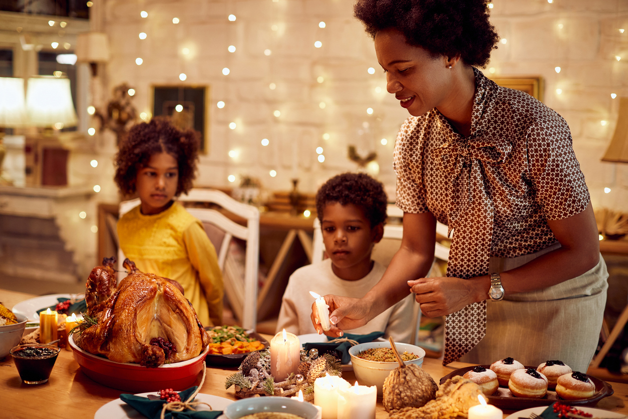 African American mother lights candles during Thanksgiving meal at dining table.