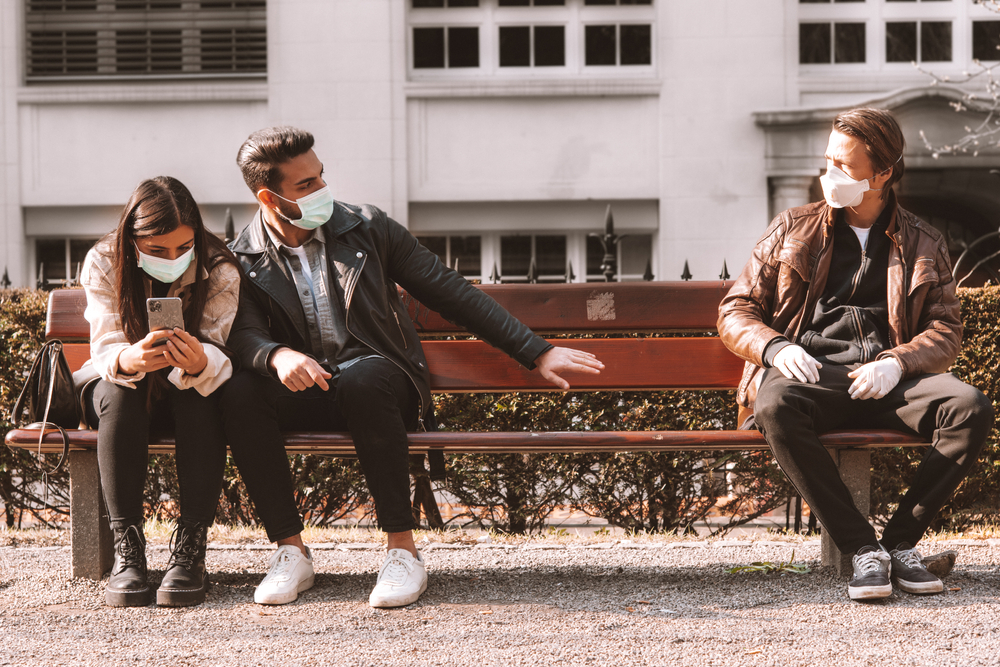 A young couple sitting on a bunch wearing face masks distances themselves from another person on the other end of the bench.